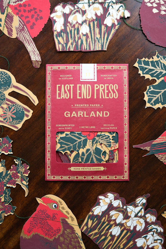 A flat-lay of a 'East End Press Garland' surrounded by various colorful paper cutouts of birds, flowers, and holly leaves.