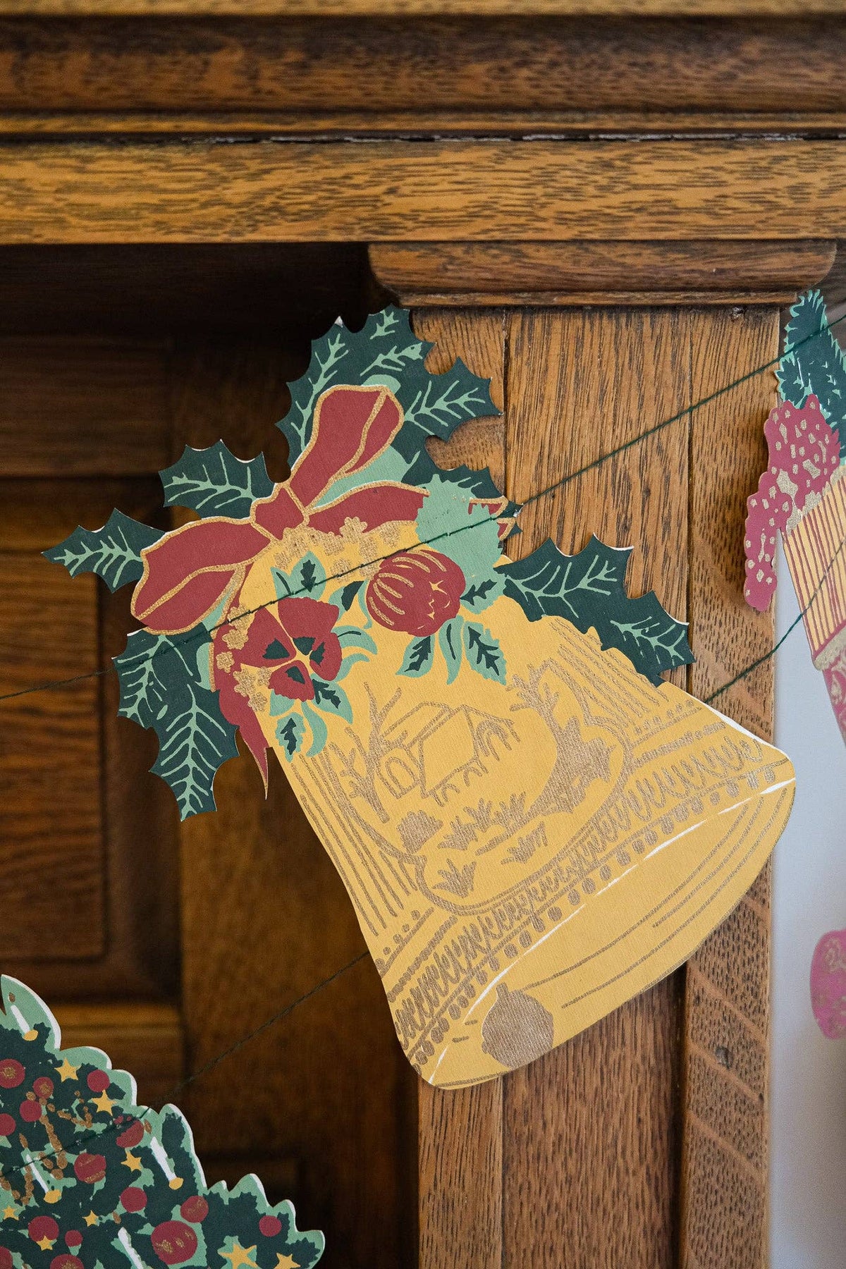 A close-up of a vintage-style Christmas garland featuring a large gold bell ornament decorated with red bows, green holly leaves, and berries, hanging against a dark wood mantel.