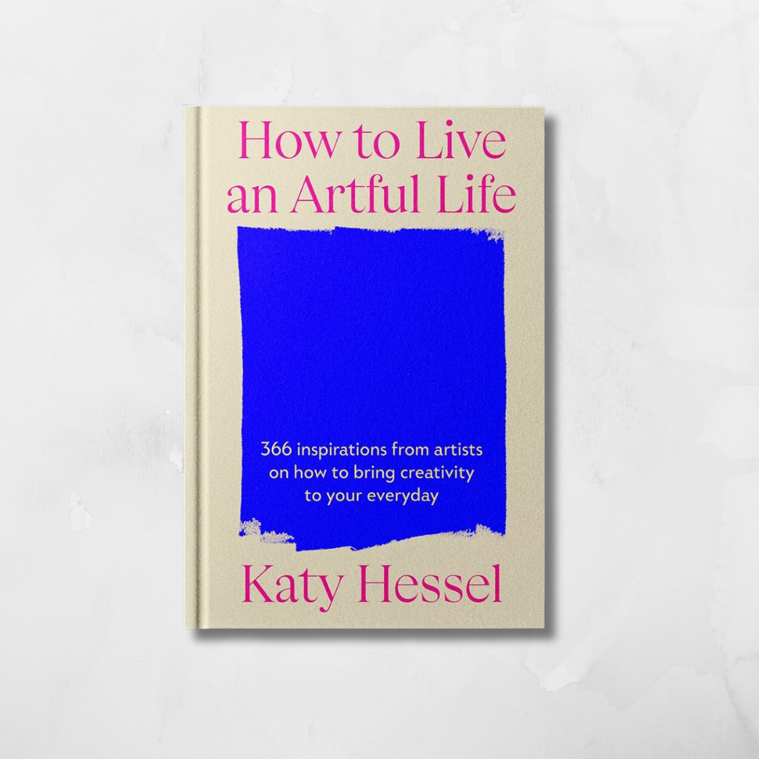 A hardcover book titled 'How to Live an Artful Life' by Katy Hessel, featuring a cream-colored cover with a large blue rectangular block in the center and pink text, set against a light gray marble background.