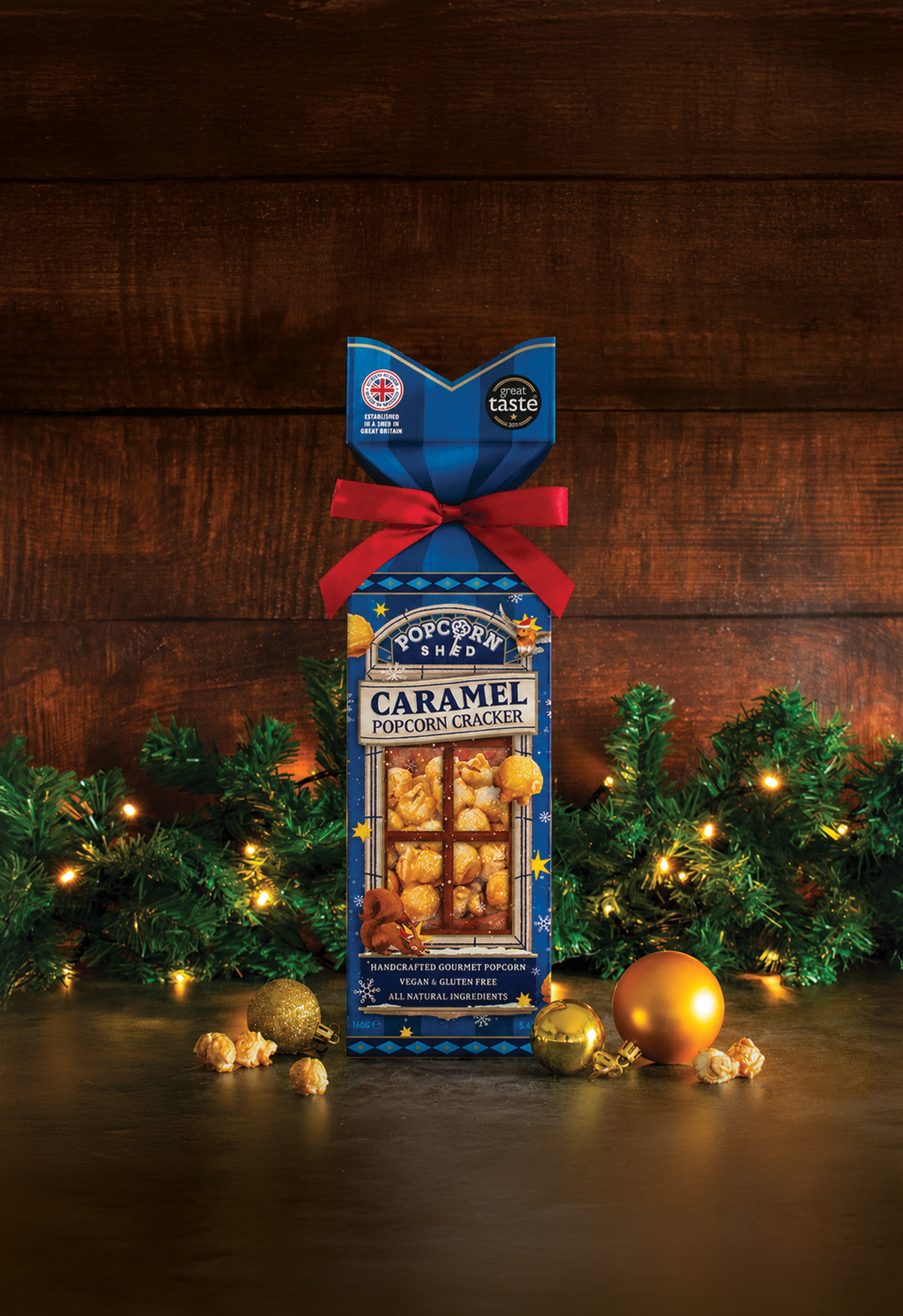 A boxed bag of Caramel Popcorn Crackers stands upright on a dark tabletop against a rustic wood plank background, decorated with green pine branches, warm string lights, gold ornaments, and scattered popcorn kernels. The package is blue with a red ribbon and a clear window showing the crackers inside.
