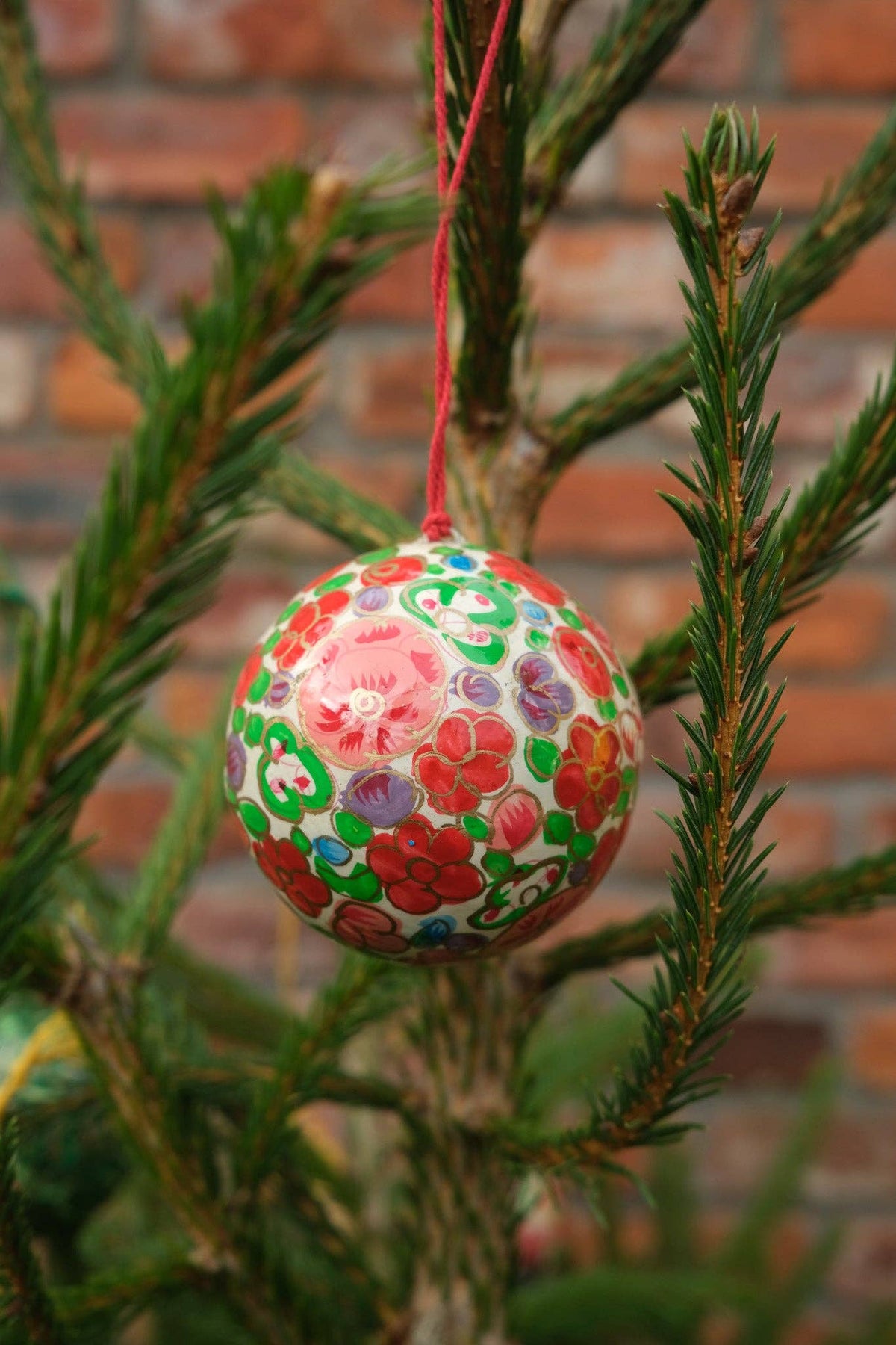 A colorful hand-painted Christmas ornament with floral patterns hangs from a red string on a green evergreen tree in front of a blurred brick wall. Made by East End Press