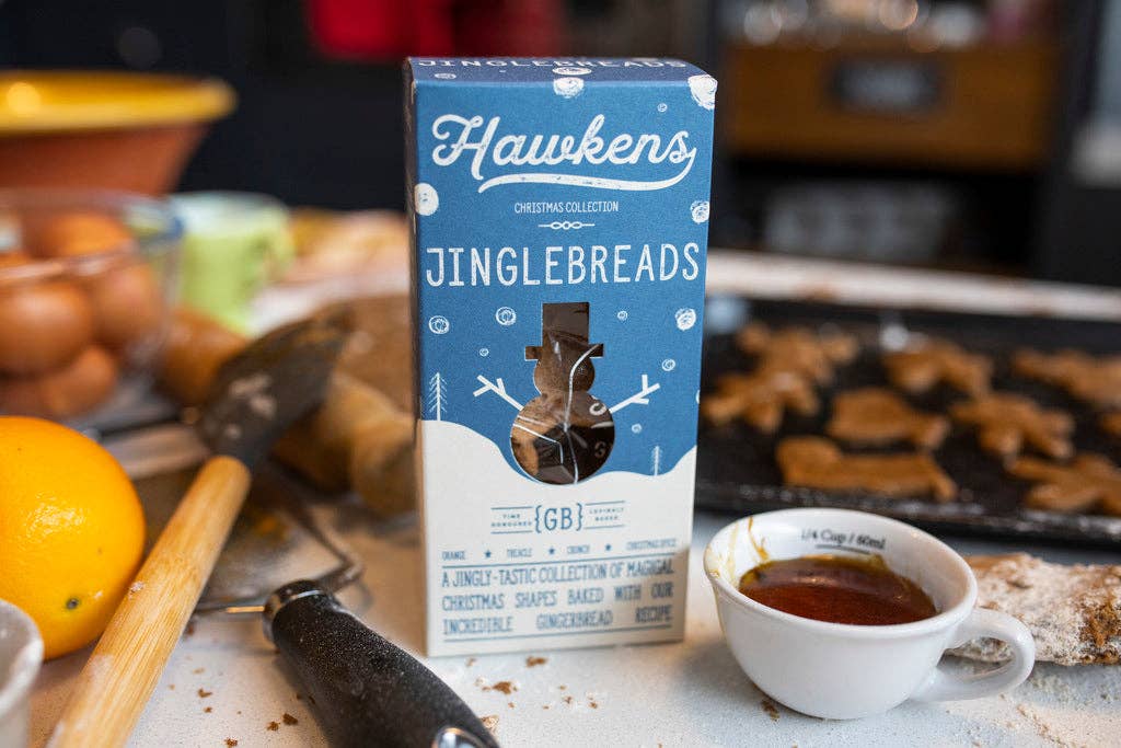 A box of Hawkens Jinglebreads centered on a kitchen counter, surrounded by baking ingredients and baked goods in a warm, blurred kitchen setting.