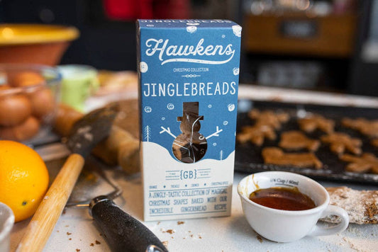 A box of Hawkens Jinglebreads centered on a kitchen counter, surrounded by baking ingredients and baked goods in a warm, blurred kitchen setting.
