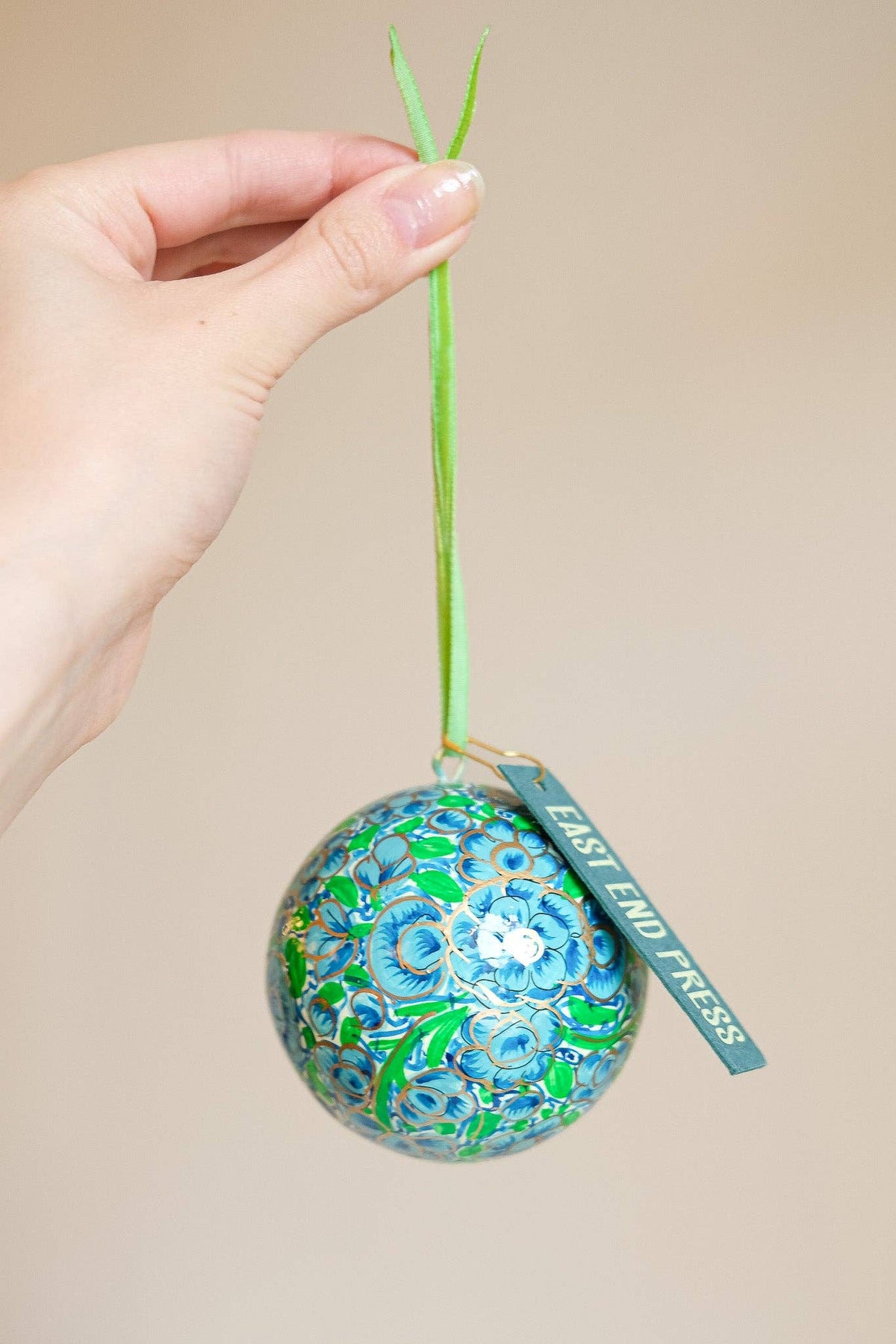 A hand holds a decorative ornament with a green stem and a hanging tag that reads 'EAST END PRESS.' The ornament is covered in a blue and green floral pattern with gold foil accents.