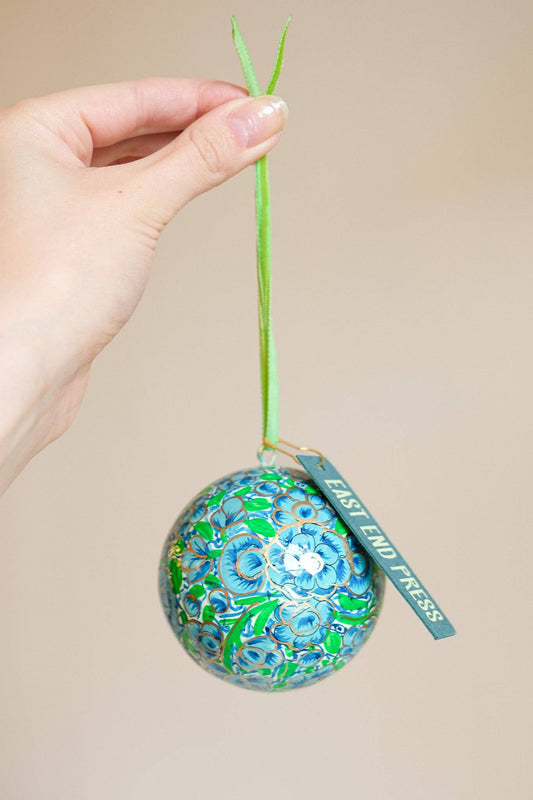 A hand holds a decorative ornament with a green stem and a hanging tag that reads 'EAST END PRESS.' The ornament is covered in a blue and green floral pattern with gold foil accents.