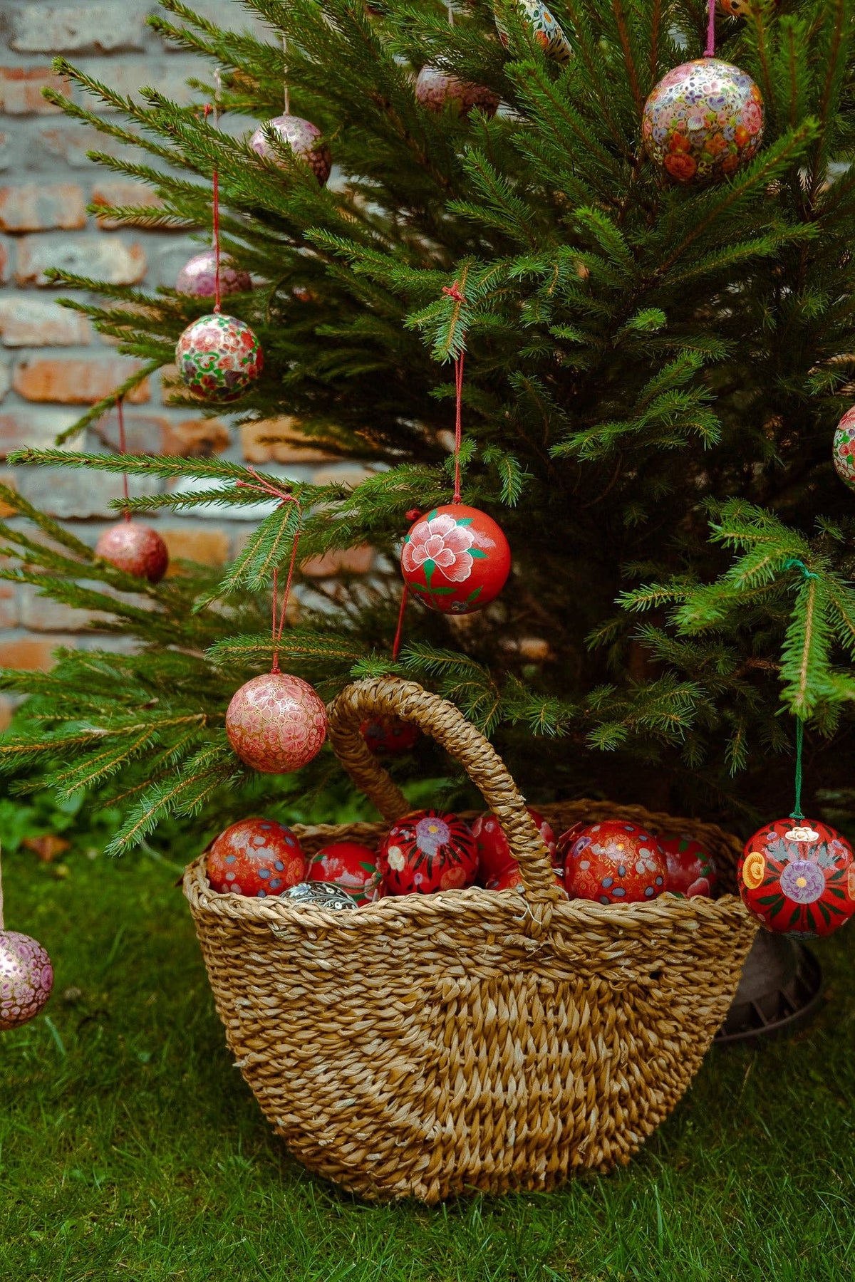 A woven straw basket filled with red and patterned Christmas ornaments sits on green grass in front of a decorated Christmas tree, with a brick wall in the background.