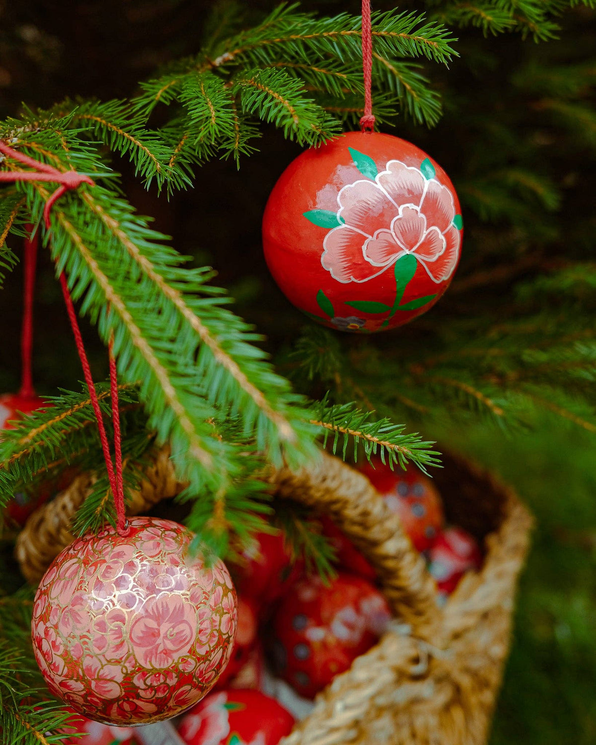 Hand-painted red East End Press Christmas ornaments hanging from green pine tree branches, with one ornament featuring a pink floral design and another with a gold floral pattern.