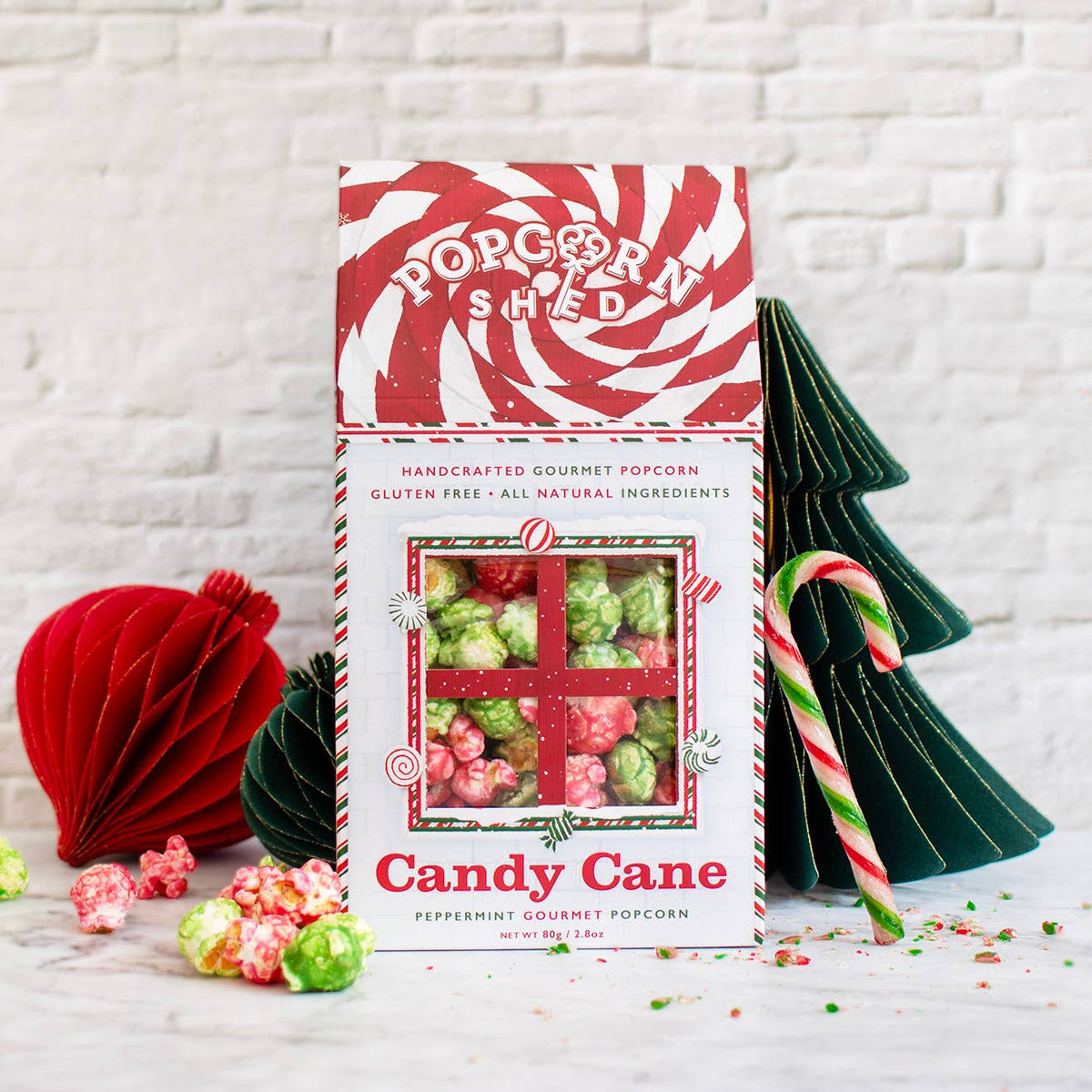 A box of Popcorn Shed Candy Cane peppermint gourmet popcorn stands on a marble surface against a white brick wall, styled with candy canes, paper ornaments, and loose popcorn pieces.
