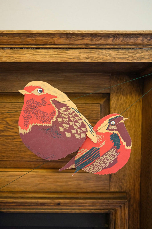 decorative bird garland shaped like red-breasted birds, hanging on a string in front of a wooden mantel or fireplace.