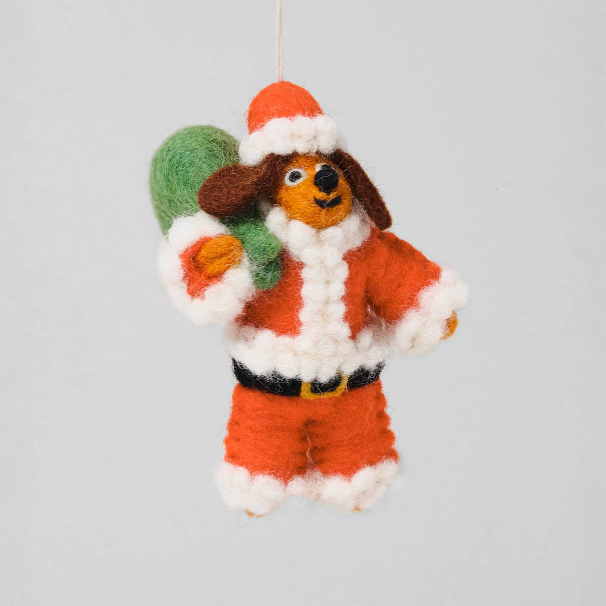 A handcrafted felt Christmas ornament shaped like a dachshund dog dressed in an orange Santa suit with white trim, holding a green sack, hanging against a light gray background.