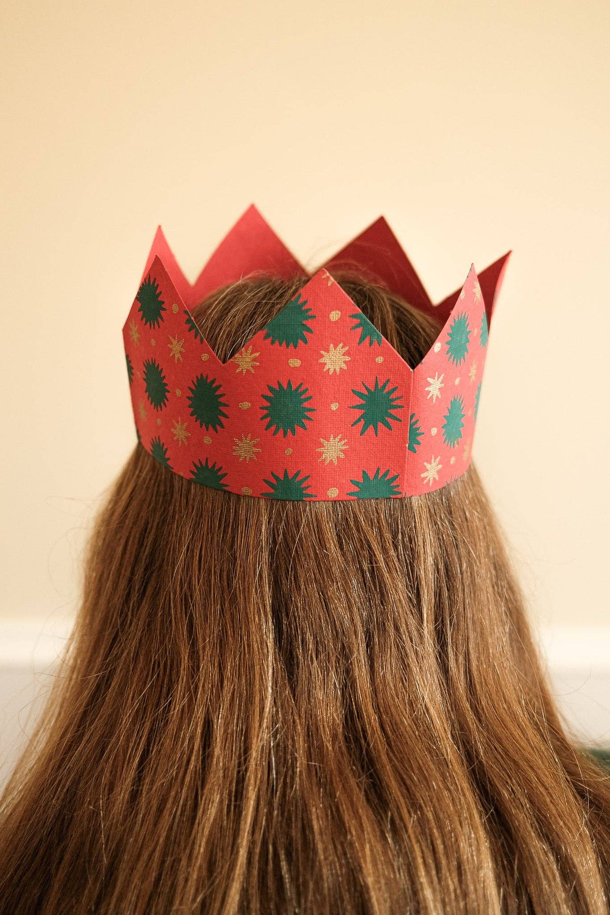 East end press red paper crown with green and gold starburst patterns worn on the head of a person with long brown hair, photographed from behind against a plain beige wall.