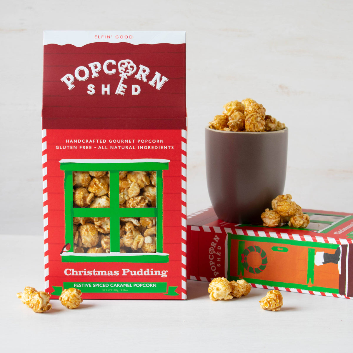 A red and green 'Popcorn Shed' box of Christmas Pudding festive speed caramel popcorn stands next to a dark brown cup filled with popcorn and a few scattered pieces on a light background.