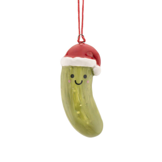 A green pickle-shaped Christmas ornament with a smiling face, wearing a red Santa hat with white trim, hanging from a red string against a white background.