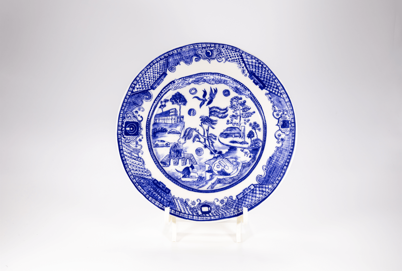 A glazed ceramic plate with a blue design featuring elements of 'normal life' such as a full English breakfast and video conferencing, displayed against a white background.